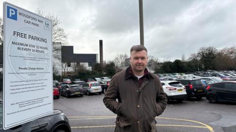 Councillor Walker stands next to the large white Woodford Way car park sign with a full car park behind him. The sign says the car park is free and that maximum stay is 12 hours. Councillor Walker where's a brown wax jacket.