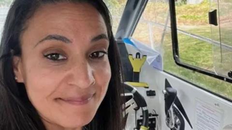 Syreeta Sandhu smiles at the camera. She is in the driver's seat of a car.