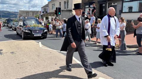 Man in black top hat and coat in front of funeral car and procession
