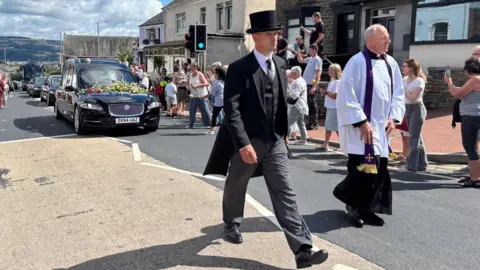 Man in black top hat and coat in front of funeral car and procession