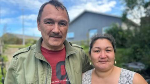 A photograph of a couple looking at the camera. The main is wearing green jacket with a red t-shirt and the woman is wearing a vest with a pattern on it. 