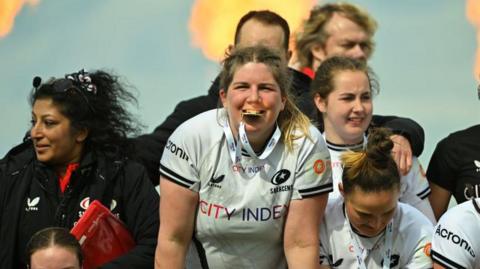 Bryony Cleall bites her PWR Cup winner's medal, surrounded by Saracens team-mates