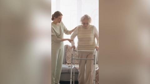A woman in scrubs helps an older woman walk with a frame