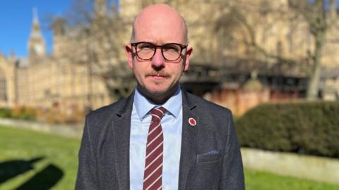 A bald man with glasses outside the Westminster Parliament on a sunny day. He is looking at the camera and is wearing a grey suit, blue shirt and maroon and grey striped tie. 