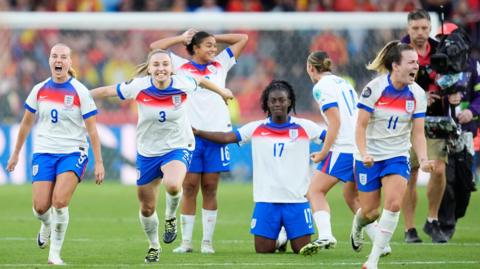 Six members of the England women's football team are celebrating on a pitch. A man with a large video camera is in the background.
