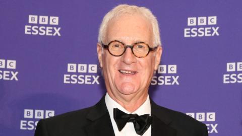 Dave Monk stands in front of a purple backdrop with several white BBC Essex logos. Dave is wearing a tuxedo jacket, white shirt and black bow tie. He has grey hair, is smiling at the camera and wears round spectacles.
