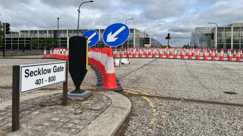 A closed road. There is a sign that reads "Secklow Gate". There are several rows of traffic cones across the road.