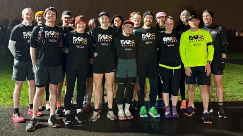 A group of runners standing together. It is raining and night time. Most of them are wearing black T-shirts which say "Run Belfast" on them in white lettering, one man to the front left of the group has the same logo but in black text on a neon yellow running top.
