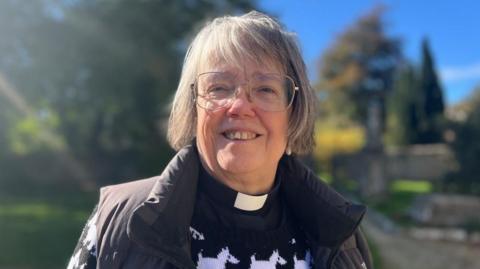 Reverend Sue Murray from the Benefice of Bisley smiles at the camera in the churchyard with the sun shining. She wears a black jumper with white dogs on it with a gilet and a clerical collar. 