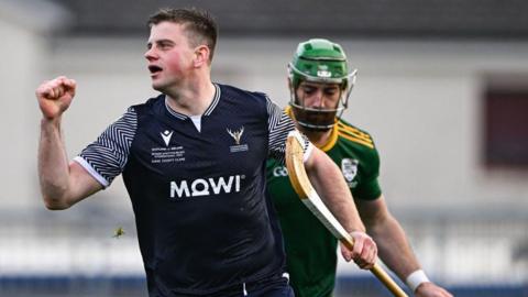 Two shinty players - one of them is in a Scotland top with a shinty stick and no helmet and dark hair and looks like he is celebrating scoring. Behind him is a player in the green and gold of Ireland with a green helmet on. 