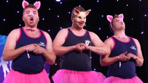 Llun yn dangos tri dyn mewn tw-tws pinc efo trwynau a chlustiau moch yn canu.