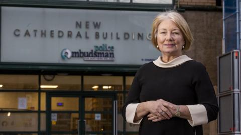 Marie Anderson standing outside the police ombudsman office in Belfast. She wears a black dress with a white collar and cuffs. She has short, blonde hair.