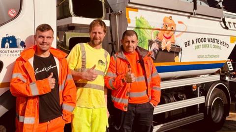 Three men in hi-viz clothing pose for a picture in front of a large food waste collection vehicle. The men are all raising their right thumb for the photo. 