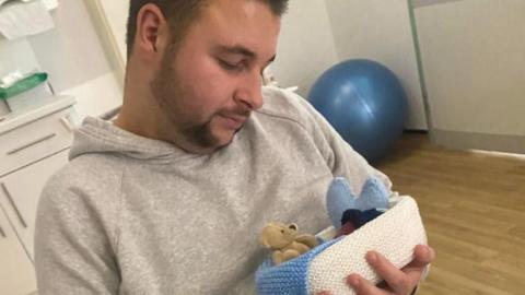A man in a grey hoodie cradles a tiny stillborn baby in a knitted blue and white basket, along with a small beige teddy and a blue felt heart.