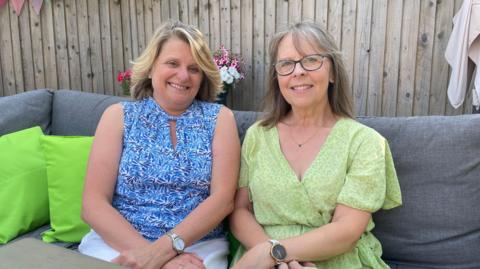 Two women sat outside of on a sofa sitting next to each other and smiling. Carole, on the left, has blonde hair and is wearing a blue and white patterned vest. Julie, on the right, is wearing a light green dress and wearing glasses.