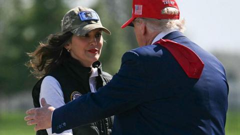 Gretchen Whitmer wears a camo baseball cap and stands as Donald Trump goes in for a hug. Trump is wearing a dark blue suit, his tie tossed over shoulder, and a red hat that reads "Trump" on the back.
