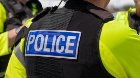 The back of a police officer. They are wearing a yellow high-vis jacket and a black vest with the word 'police' across the back. 