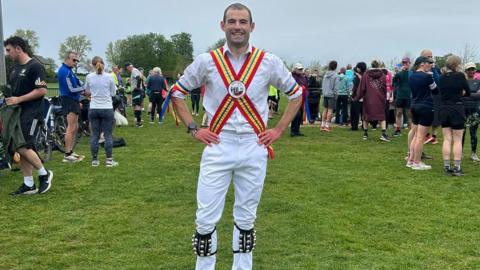 A man with short brown hair in a field dressed in Morris dancing garb.