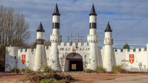 The entrance to the abandoned Camelot theme park. The image shows a mock white castle frontage with a barbican and turrets. There are coats of arms on the walls and overgrowth in the foreground. 