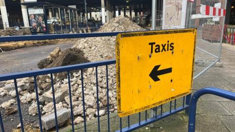 A sign attached onto fencing at Great Yarmouth Market Gates bus station on Temple Road, directs passengers to the nearest taxi ranks: Theatre Plain (outside Costa Coffee) and Market Gates Road. Behind the fencing is a pile of rubble on the roadway and the Market Gates shopping centre.