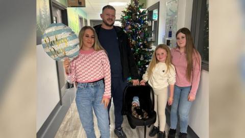 A woman wearing a pink jumper and holding a balloon saying 'baby boy' standing in a hospital corridor with her partner, a man in a black hoodie, and two daughters. Her partner is carrying a baby carrier with a baby inside wearing a blue bobble hat. 