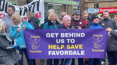 About a dozen people stand behind a purple banner saying "Get behind us to help save Favordale"