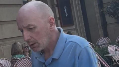 Stewart Irwin pictured outside a cafe seating area. He has short stubble and is bald. He is wearing a blue button collar t-shirt and is looking down.