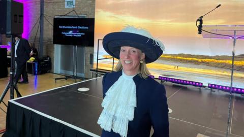 A woman with blonde hair wearing a wide-brimmed blue hat with white feathers and a blue jacket with a large white lace ruffle