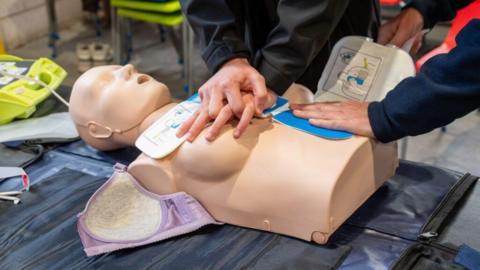 A female CPR dummy, on a surface, with a bra around it, and three hands on it, there is equipment all around it. You can see three arms. 
