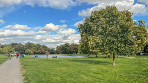 File photo of Clapham Common in the daytime. There is green grass, some trees and people walking on a path and sitting on a bench looking at a pond.