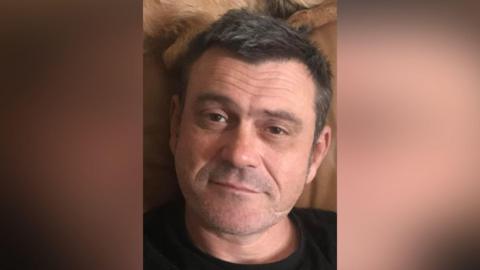 Peter Graham has short black hair and stubble. He is looking at the camera and has a black T-shirt. 