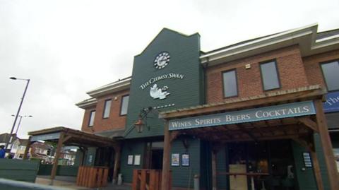 External shot of a brick-built pub with a large green panel featuring a clock and The Clumsy Swan pub sign.