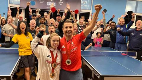 Paralympian medallists Bly Twomey and Will Bayley wear their medals and punch their fists in the air in front of a crowd at Brighton Table Tennis Club