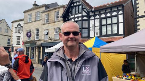 Image of man in the centre of the picture wearing sunglasses, in a purple raincoat who has short white hair standing in the middle of a street with stalls to the left of him and people to his right