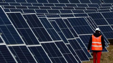 Solar farm panels lie at an angle on grassland, pointing towards the sky. In front of them, a man in an orange hi-vis jacket, white hard hat and red trousers is walking away from the camera.