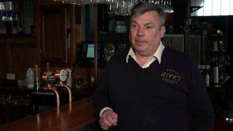 a man in a black jumper and white shirt looks serious as he leans against the bar of a dimly lit restaurant