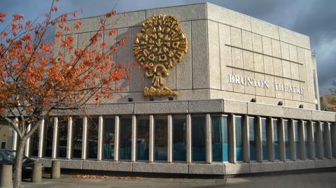 A side view of the exterior of Brunton Theatre. The building is grey, with a gold design on one side. The letters 'Brunton Theatre' are on the other side in silver. A tree with red and orange leaves is in front of the building above three stone bollards.