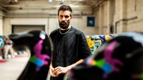 Serge Pizzorno wearing a black t shirt and chain, stood among colourfully painted elephant sculptures