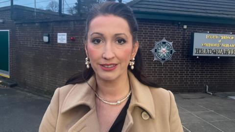 Emma Little-Pengelly who has long dark hear and is wearing a tan-coloured coat coat and pearl earrings and necklace. She is standing in front of the brown brick wall at the front of the Police Service of Northern Ireland headquarters.