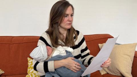 Beth Kojder holding her newborn baby and reading through an energy bill