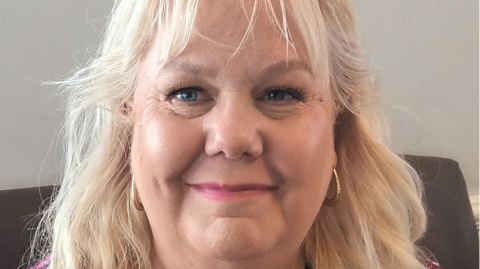 A woman in her 50s with long blonde hair and blue eyes. She is wearing gold hooped hearings and is smiling.