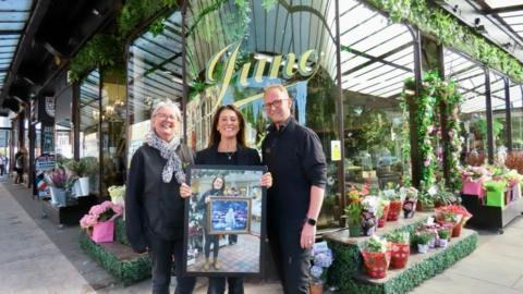Liz (centre) holds a framed picture of a picture of her holding a picture of Bessie.  Members of staff Pauline (left) and Matthew stand either side of her. They are in front of the shop  
