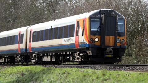 A South Western Railway train makes its way along the railway line near to Basingstoke in Hampshire. Green grass is seen either side and tall trees in the background.