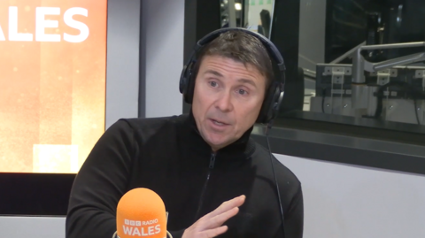 Dean Harris who has dark hair and eyes and is wearing a black zipped up fleece, sat in a radio studio