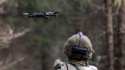 The back of a soldiers head, wearing a helmet as the soldier looks out at a drone flying ahead of them.