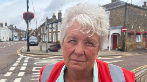 A lady with short white/grey hair swept across her forehead, she is wearing a high-viz orange and grey tabard over a green top and black blouse. Behind her is a street with cars and buildings with flowers visible on hanging baskets and planters.