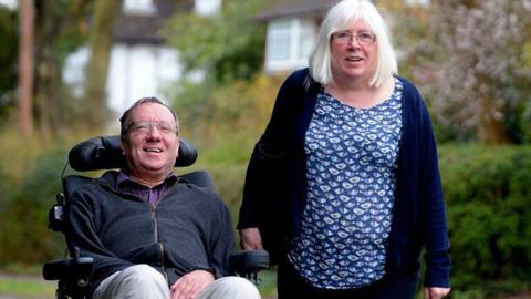 A man (David Setters) in a wheelchair alongside his wife who is his principal carer,  in a park. They are both smiling. There are trees and hedges behind them.