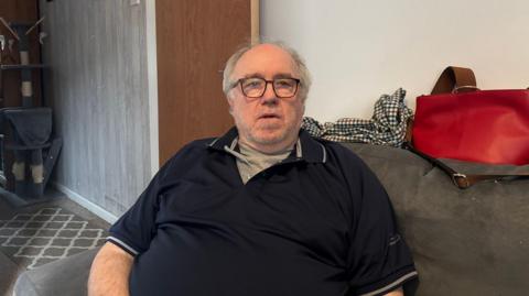 A man is sitting on a grey sofa. He has grey hair and back glasses and grey stubble. He is wearing a black t-shirt