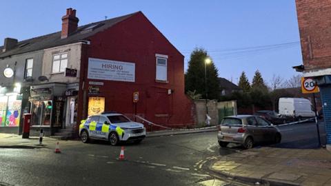 A police car is parked on a quiet street in front of a row of brick shops and flats during early evening light. Traffic cones surround the vehicle and blue and white police tape cordons off the scene.