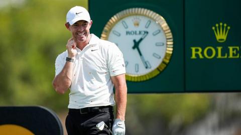 Rory McIlroy looks on after firing his tee-shot at the 14th hole of the Arnold Palmer Invitational. He is wearing a white cap, white Nike polo shirt and black trousers. He is holding his club in his gloved left hand. A large Rolex clock and advertising board can be seen behind him